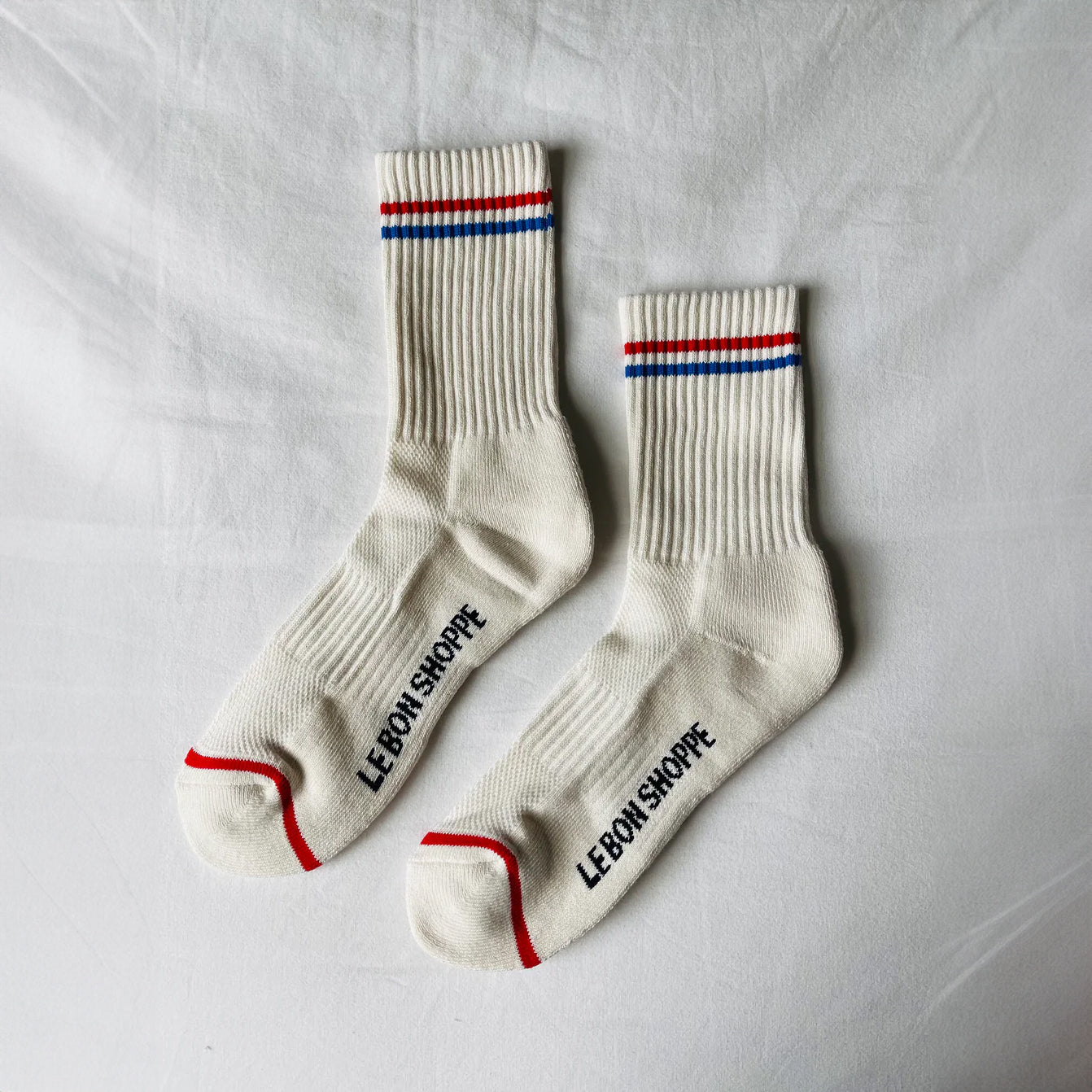 Close-up of cream Le Bon Shoppe Boyfriend Socks with red and blue striped cuffs and logo detail on the sole.