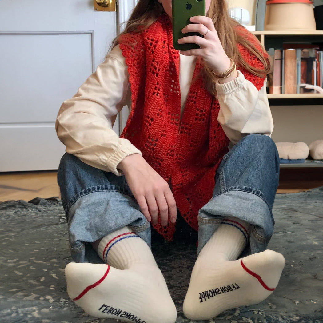 Person wearing Le Bon Shoppe Boyfriend Socks with red and blue stripes, sitting cross-legged in denim jeans and red knit vest.