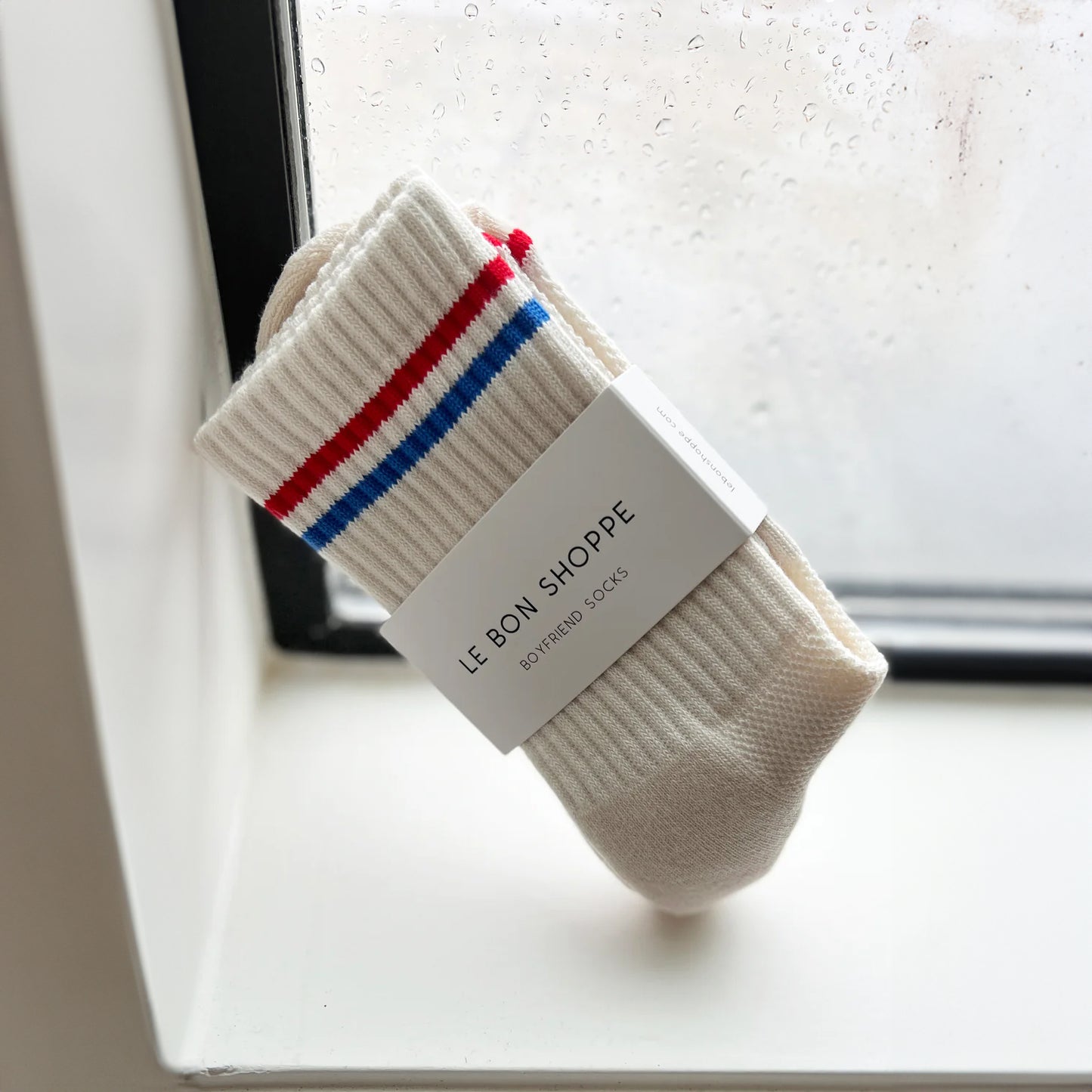 Pair of cream Le Bon Shoppe Boyfriend Socks neatly rolled with branded wrap label, placed on a windowsill.