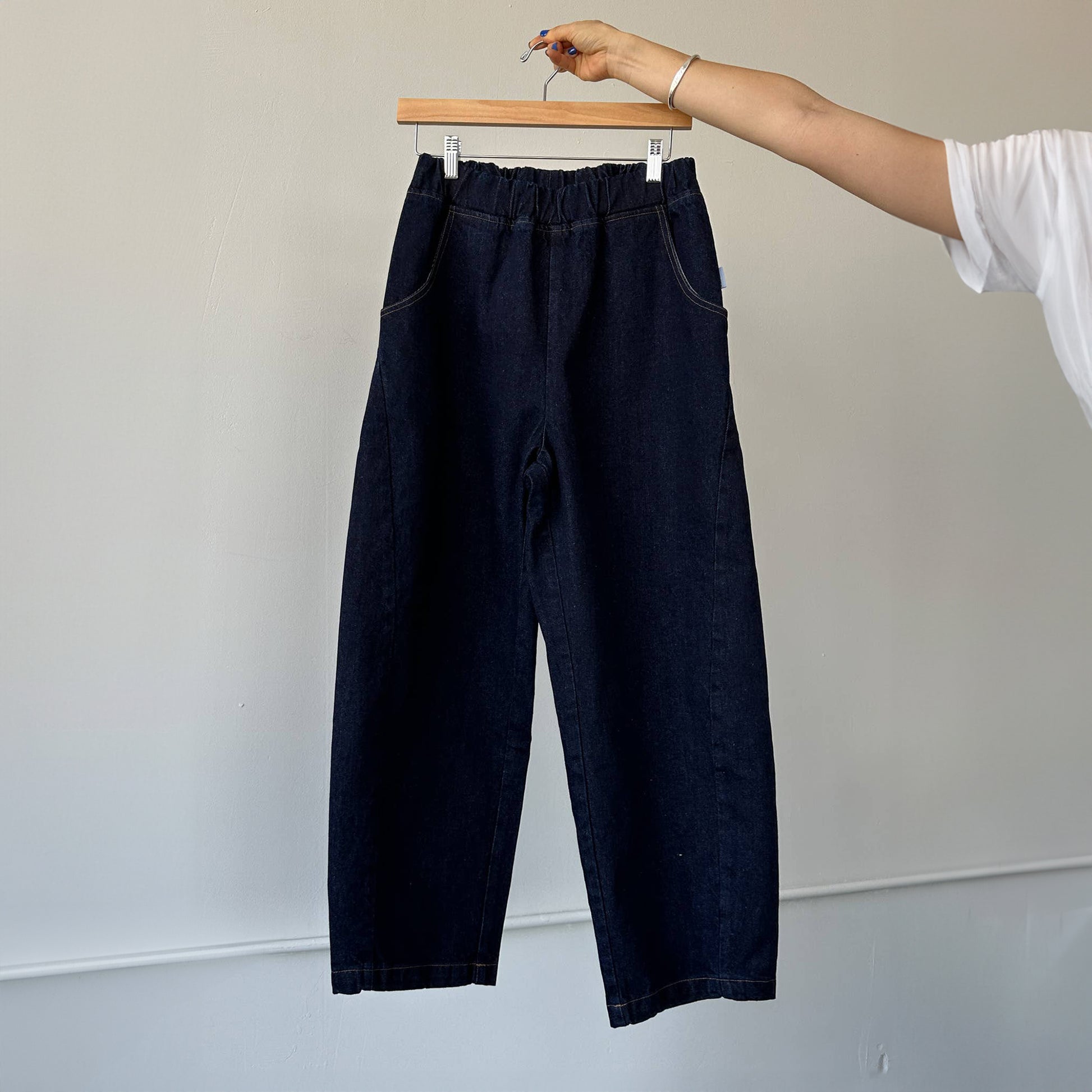 Hand holding the Arc Pants in Raw Denim – The jeans are suspended from a hanger, showcasing their structured yet soft silhouette and deep pockets.