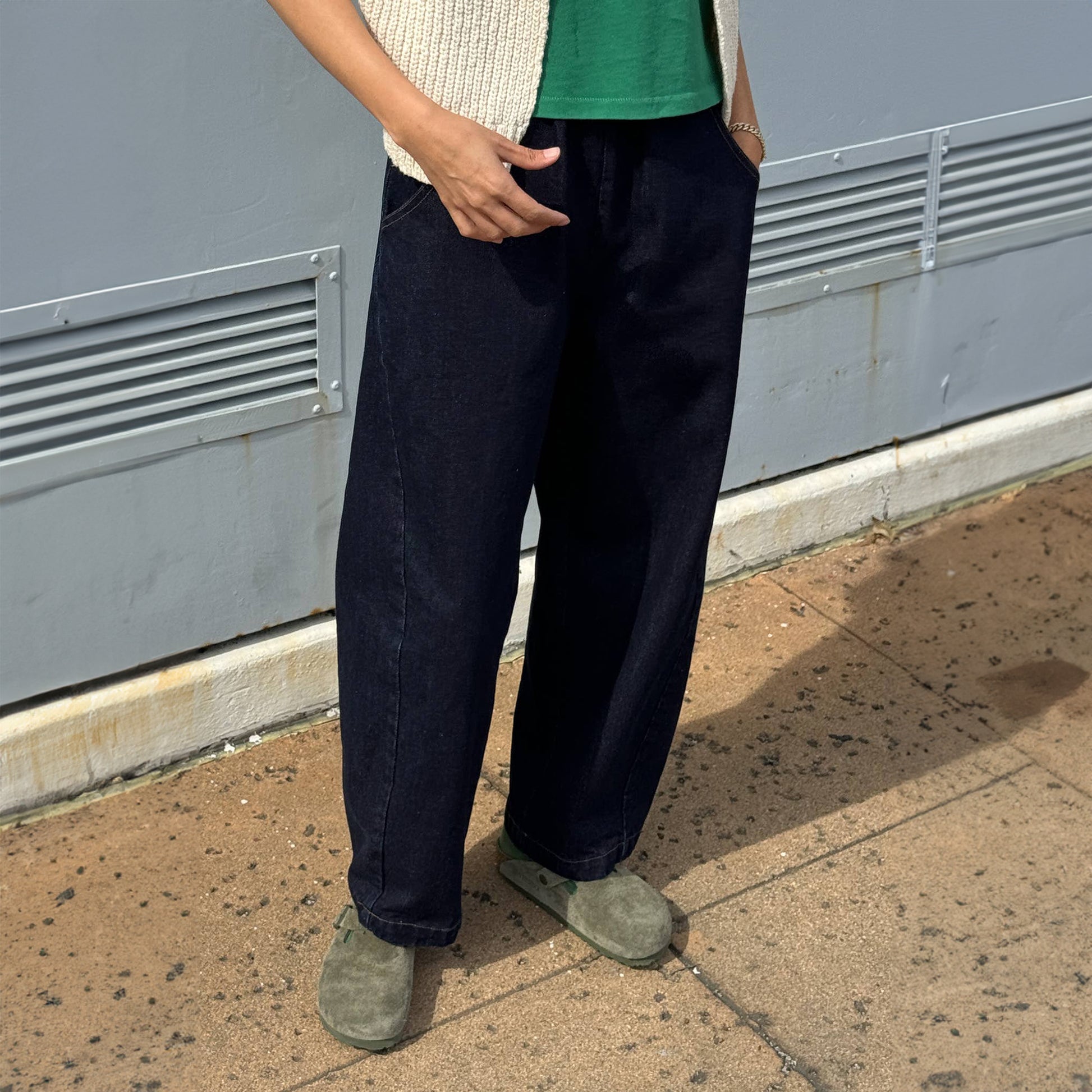 Front view of Arc Pants in Raw Denim outdoors – Styled with a green top and cream knit vest, with hands tucked into the deep pockets, showing the ease of wear.