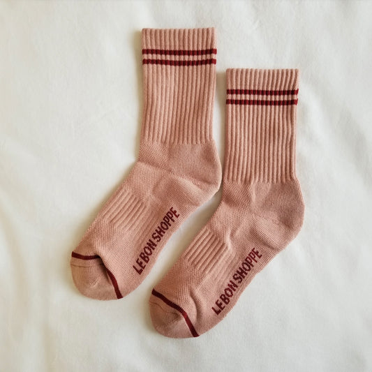A pair of vintage pink ribbed crew socks with dark red stripe details, laid flat on a white fabric background, showing the Le Bon Shoppe branding on the sole and contrast toe accents.