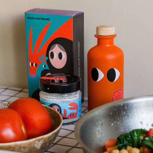 Table with tomatoes, a jar, an orange bottle, and a bowl of salad with a colorful box in the background.