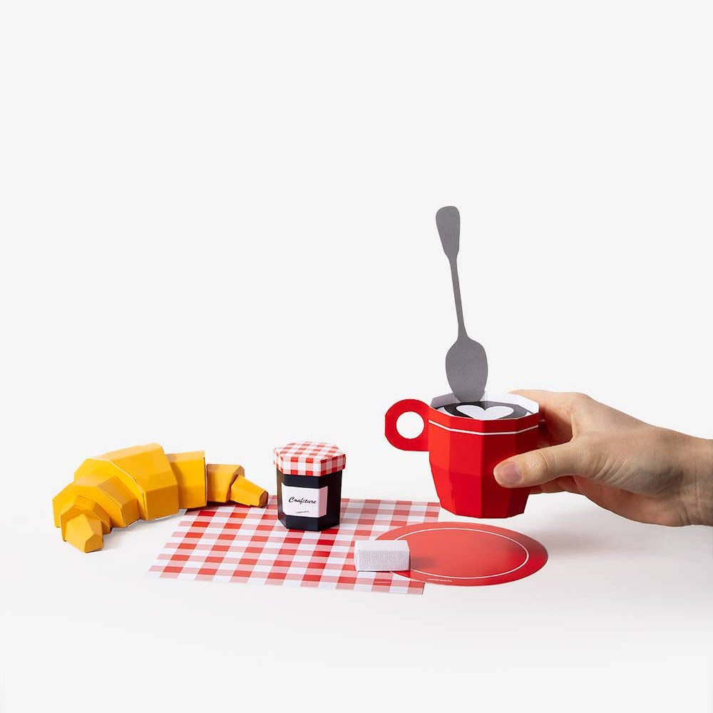 Red mug with a spoon on a checkered tablecloth, accompanied by a croissant and jam.