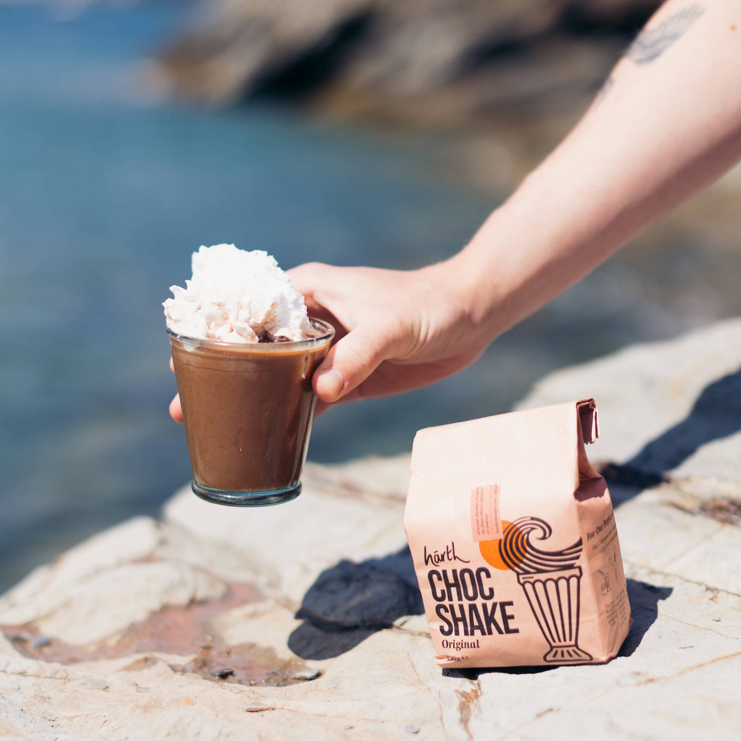 Hand holding a Choc Shake with whipped cream by a scenic coastal background