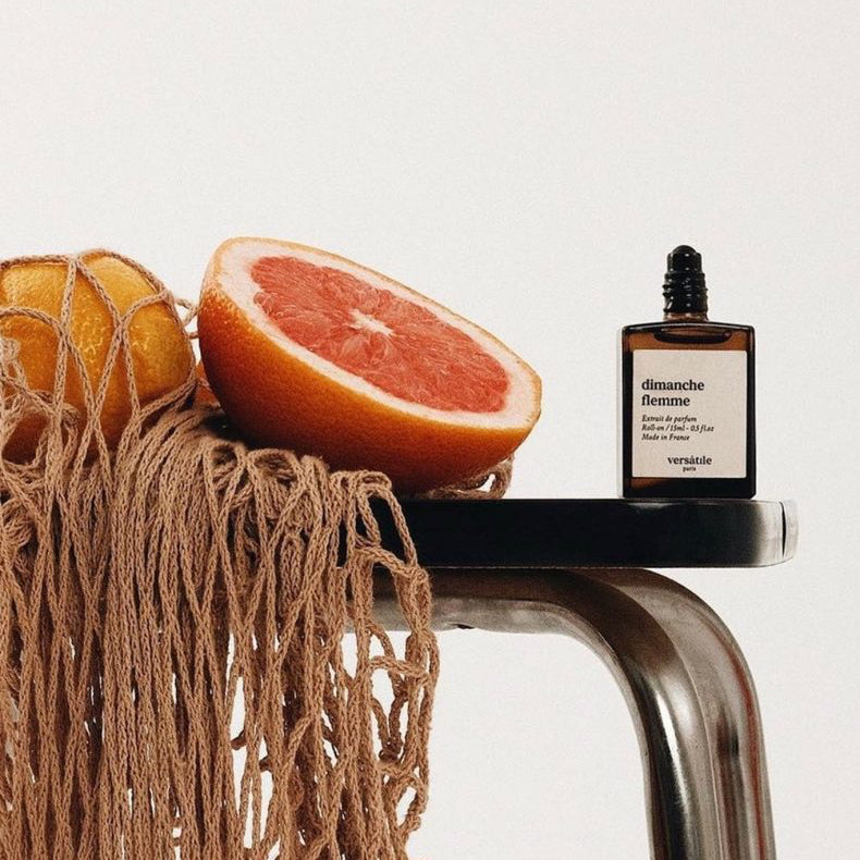 Bottle of Aromachologie perfume on a metallic stand with a grapefruit and a woven bag on a light background