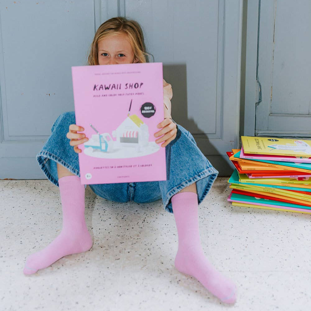 Child holding a pink book titled 'Kawaii Shop' in front of a stack of books on a light-colored floor.