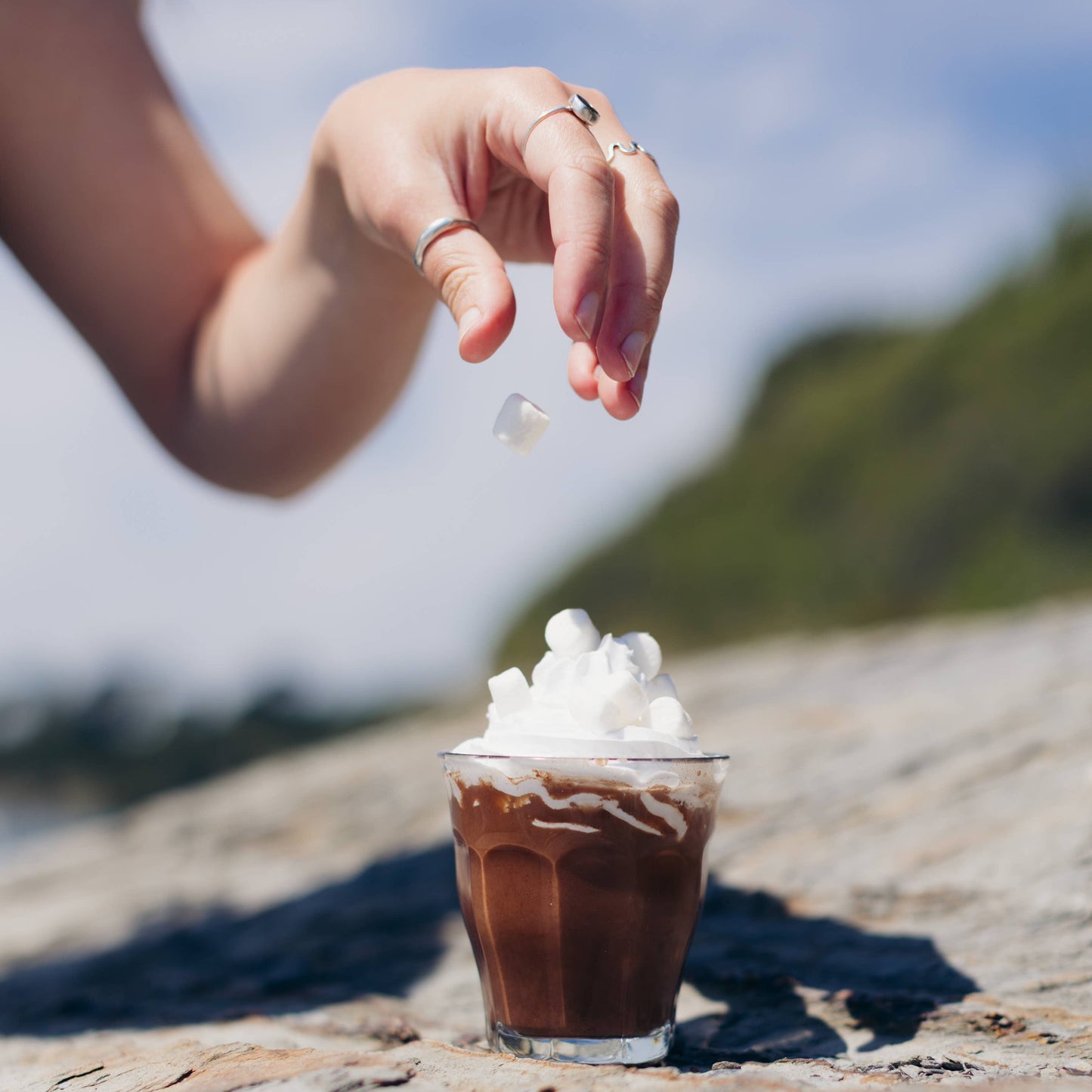 Hand sprinkling mini marshmallows onto a small glass of chocolate milkshake with whipped cream on a stone surface.