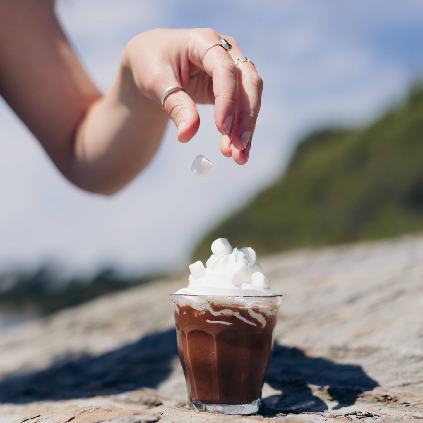 Hand sprinkling mini marshmallows onto a small glass of chocolate milkshake with whipped cream on a stone surface.