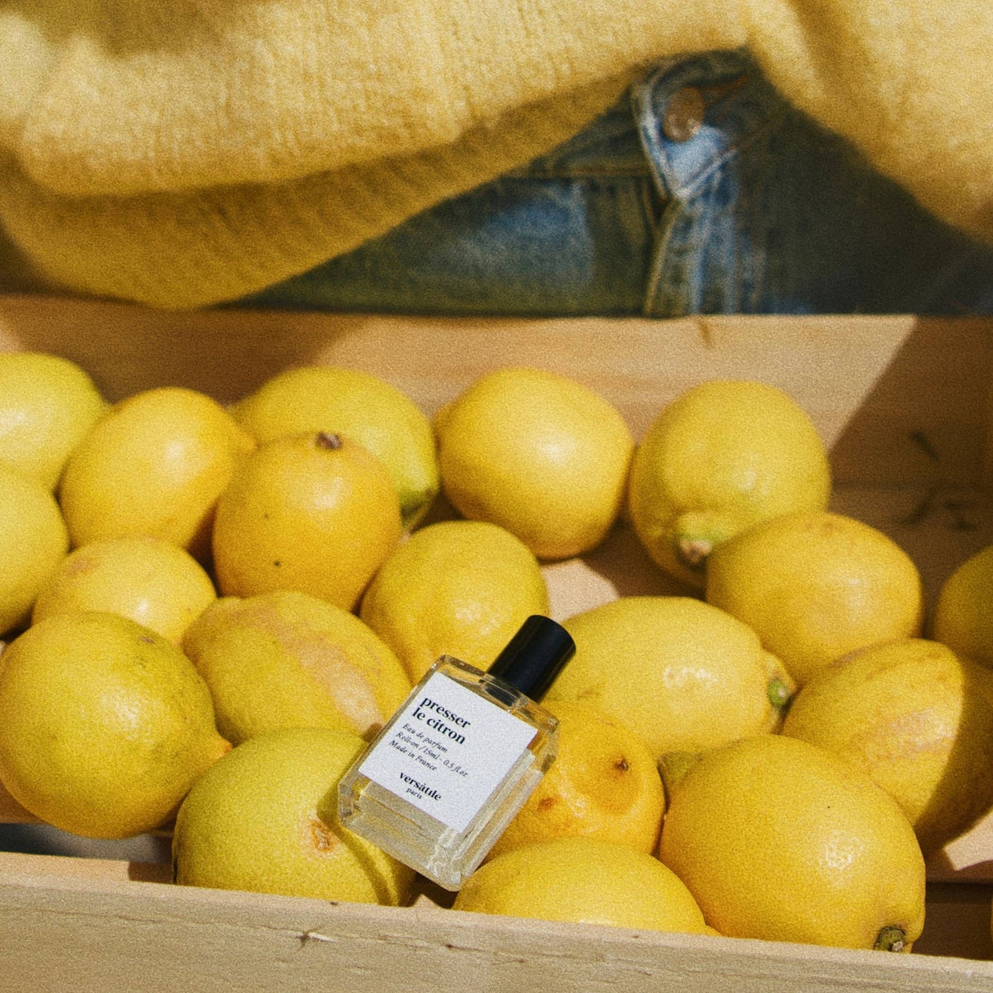 A bottle of 'Squeeze the Lemon - Eau de Parfum' placed among fresh lemons displayed in a wooden box, with a yellow cardigan in the background.