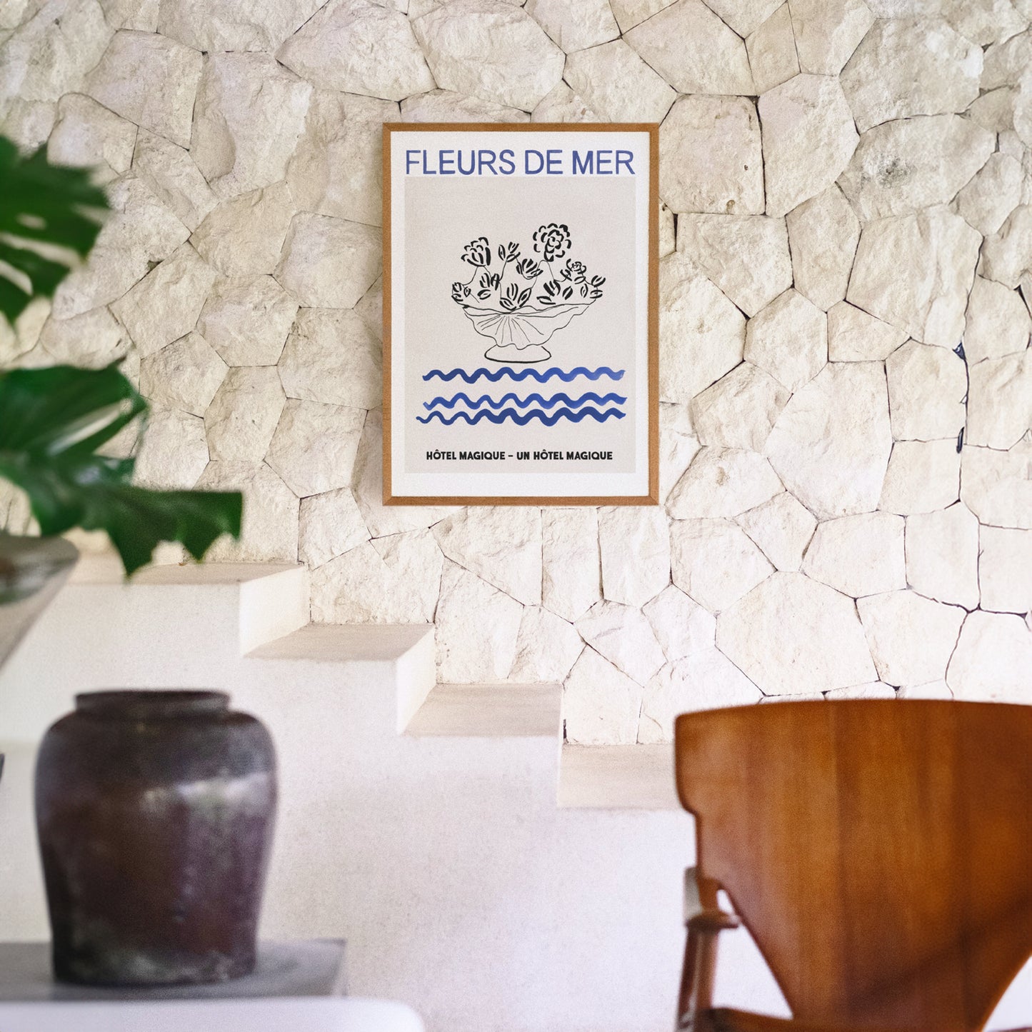 Hotel Magique Fleurs De Mers Print. The background is beige. The flowers and the flower pot is black. The waves below are dark blue in colour. “Fleurs De Mer” is written in dark blue. The print is framed and hung on a stone wall in a brightly lit room, a pot and chair in the foreground.