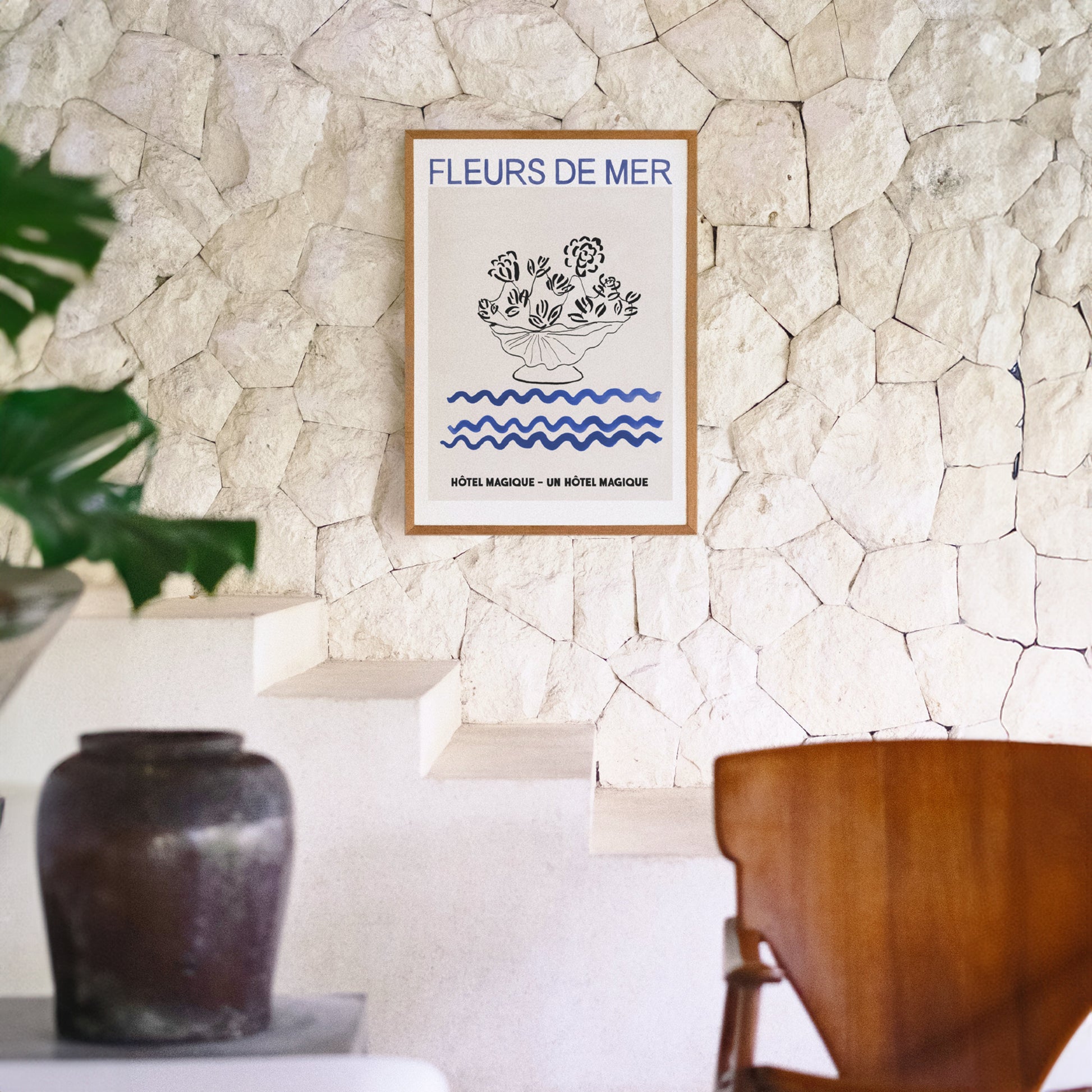 Hotel Magique Fleurs De Mers Print. The background is beige. The flowers and the flower pot is black. The waves below are dark blue in colour. “Fleurs De Mer” is written in dark blue. The print is framed and hung on a stone wall in a brightly lit room, a pot and chair in the foreground.