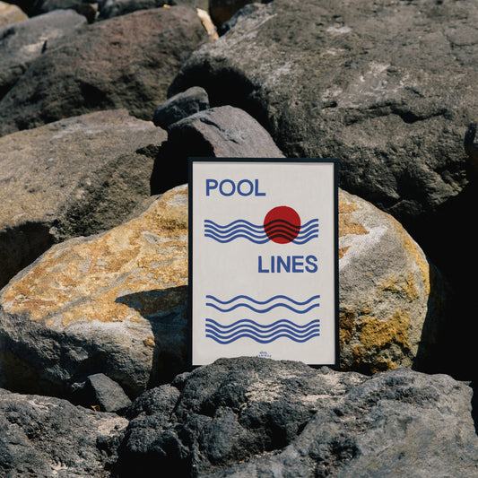 A Hotel Magique Pool Lines A3 Print. The print has a white background with Pool lines written in caps in dark blue, the waves are dark blue as well. The circle is red. The frame is placed on grey rocks
