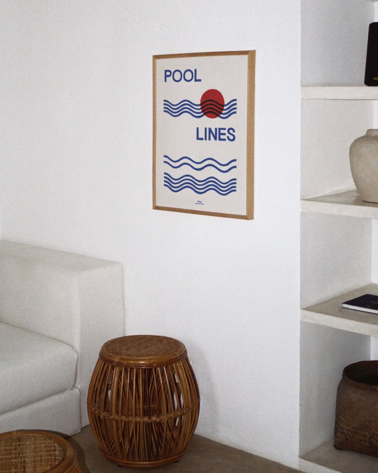 An image depicting Hotel Magique Pool Lines A3 Print. The print has Pool lines written in caps in dark blue, the waves are dark blue as well. The circle is red. The background is white. The print is framed with beige border. The frame is hung on a white wall in a room next to a sofa and other house decor.