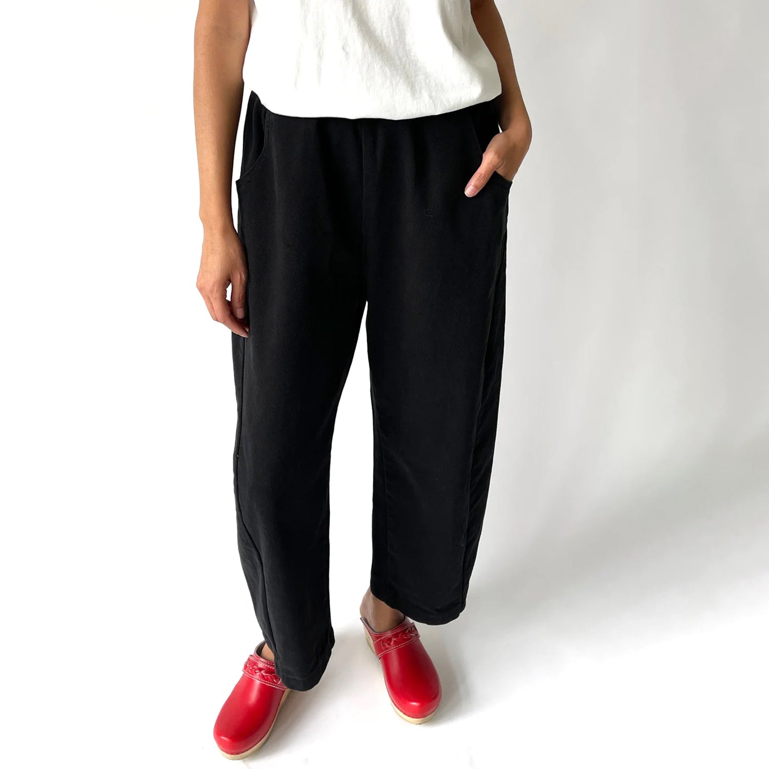 Model wearing Le Bon Shoppe Arc Pants in black, featuring a relaxed cropped fit, curved leg, and elastic waistband. Styled with red clogs and a white tee against a plain background.