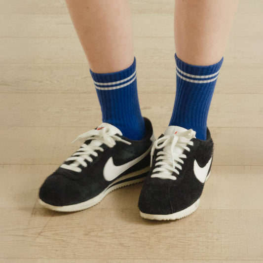 Close up of a person wearing cobalt blue ribbed crew socks with black shoes standing on beige wood flooring