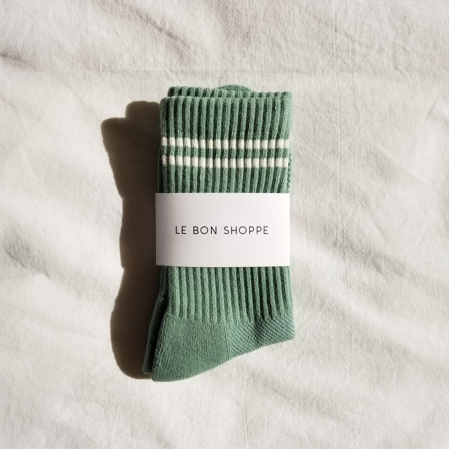 A pair of meadow green ribbed crew socks with white stripes near the cuff, neatly folded and wrapped with a white Le Bon Shoppe label, placed on a white fabric background.