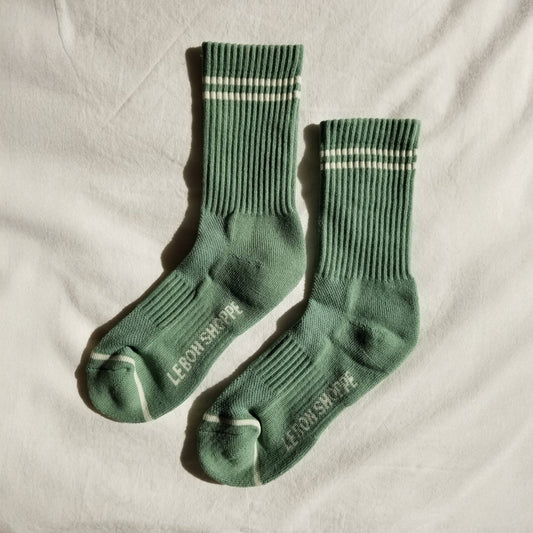 A pair of meadow green ribbed crew socks with white stripe details, laid flat on a white fabric background, showing the Le Bon Shoppe branding on the sole and contrast toe accents.