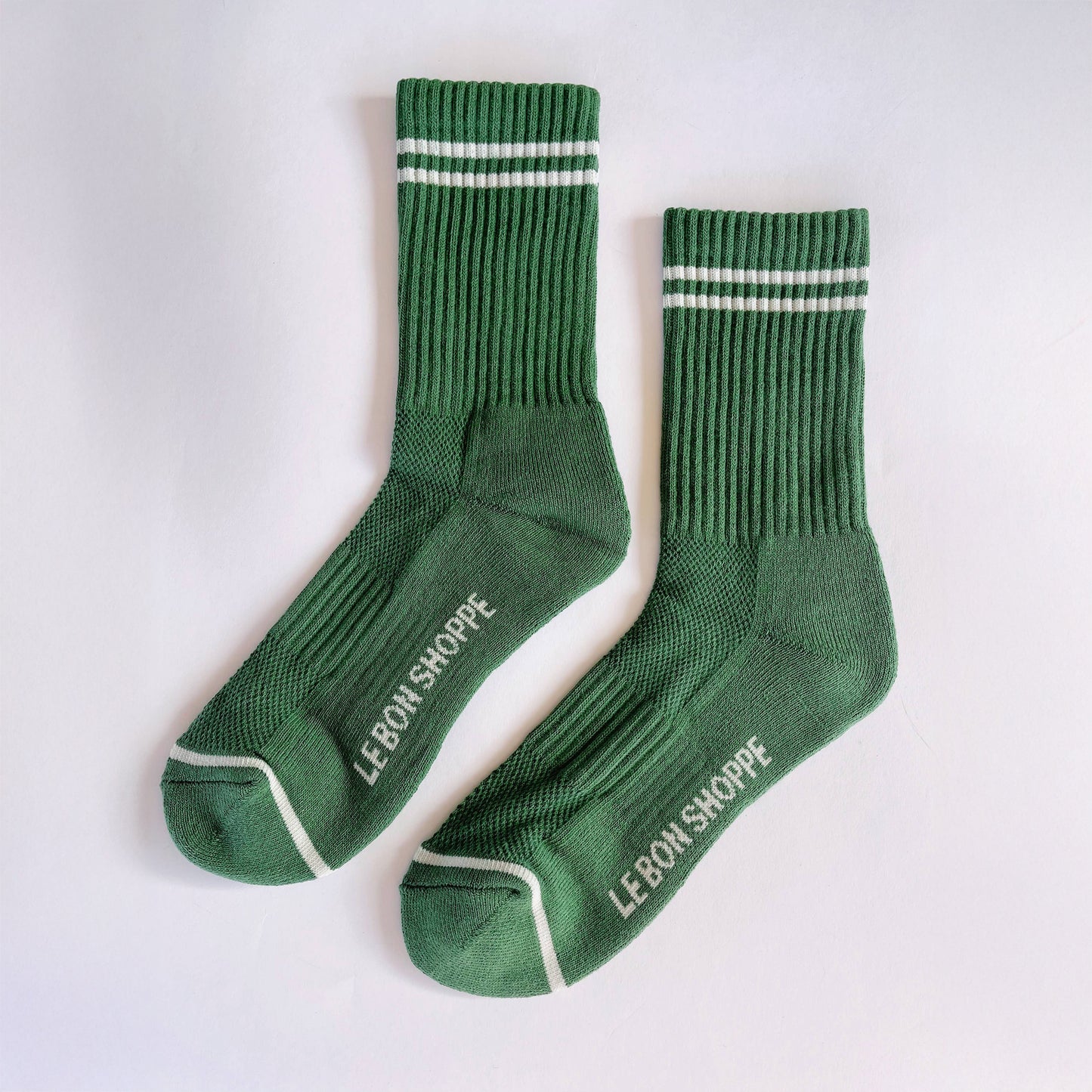 A pair of green moss coloured ribbed crew socks with white stripe details, laid flat on a white fabric background, showing the Le Bon Shoppe branding on the sole and contrast toe accents.