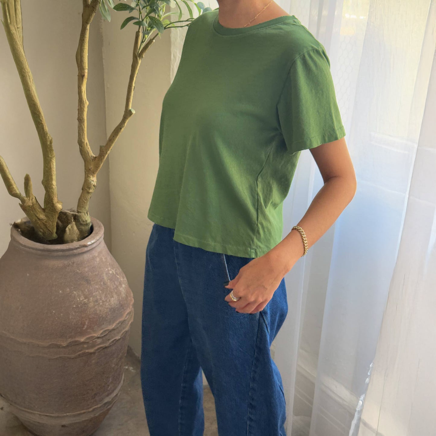Person wearing a green t-shirt and blue jeans standing next to a potted plant indoors.