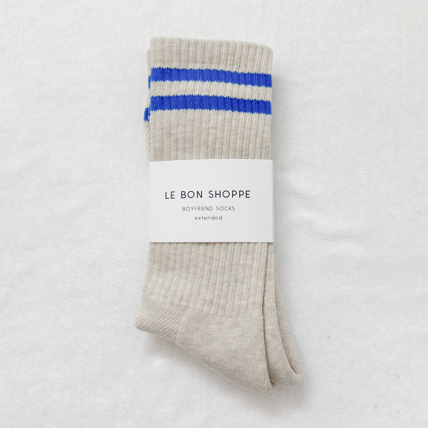 A pair of Le Bon Shoppe Extended Boyfriend Socks in an icy off-white shade with blue stripe details, neatly folded with the brand’s signature white packaging label against a white background.