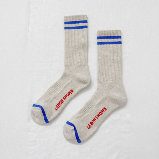 A pair of Le Bon Shoppe Extended Boyfriend Socks in an icy off-white shade with blue stripe details, laid flat against a white surface, showcasing the "Le Bon Shoppe" text on the soles and the blue accents on the cuffs and toes.
