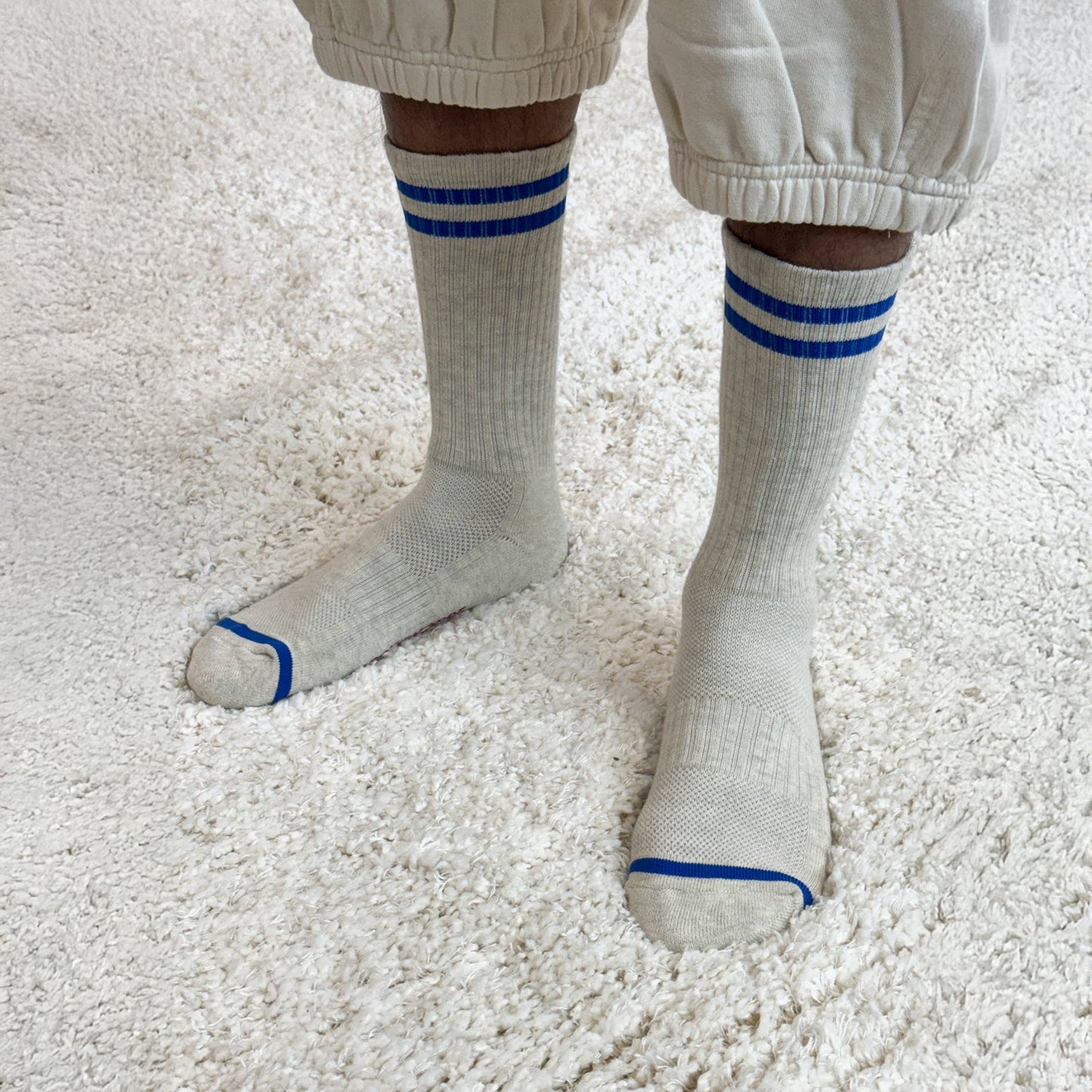 A person wearing the Le Bon Shoppe Extended Boyfriend Socks in Ice while standing on a plush, cream-coloured rug. The ribbed texture and blue stripe details are visible, paired with natural-toned balloon pants.
