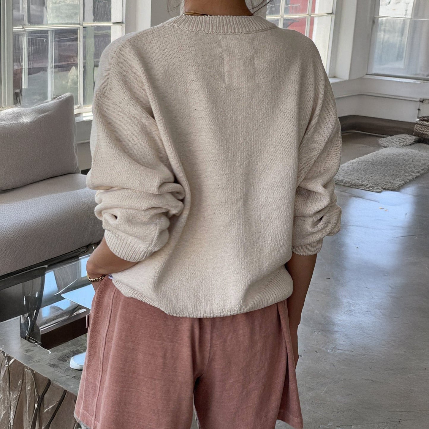 Person wearing a naturel sweater and pink pants in a room with a couch and windows.