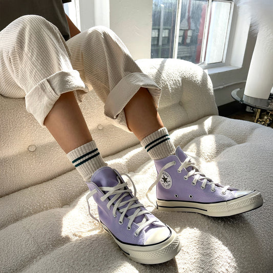 A person wearing oatmeal coloured ribbed socks with dark green stripes near the cuff with purple shoes sitting on a white couch
