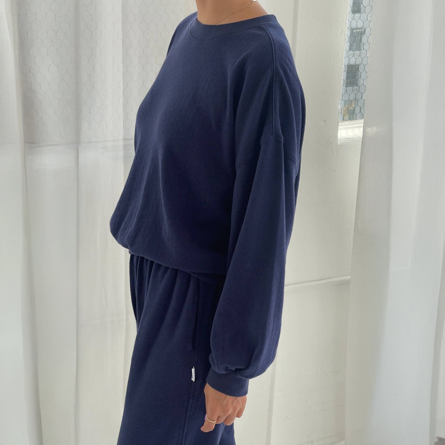 Person wearing a navy blue sweatshirt and pants in a minimalistic room with white curtains.