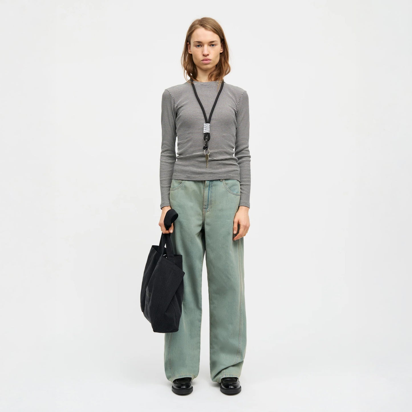 Person wearing a grey long-sleeve shirt and light blue jeans, holding a black bag on a white background