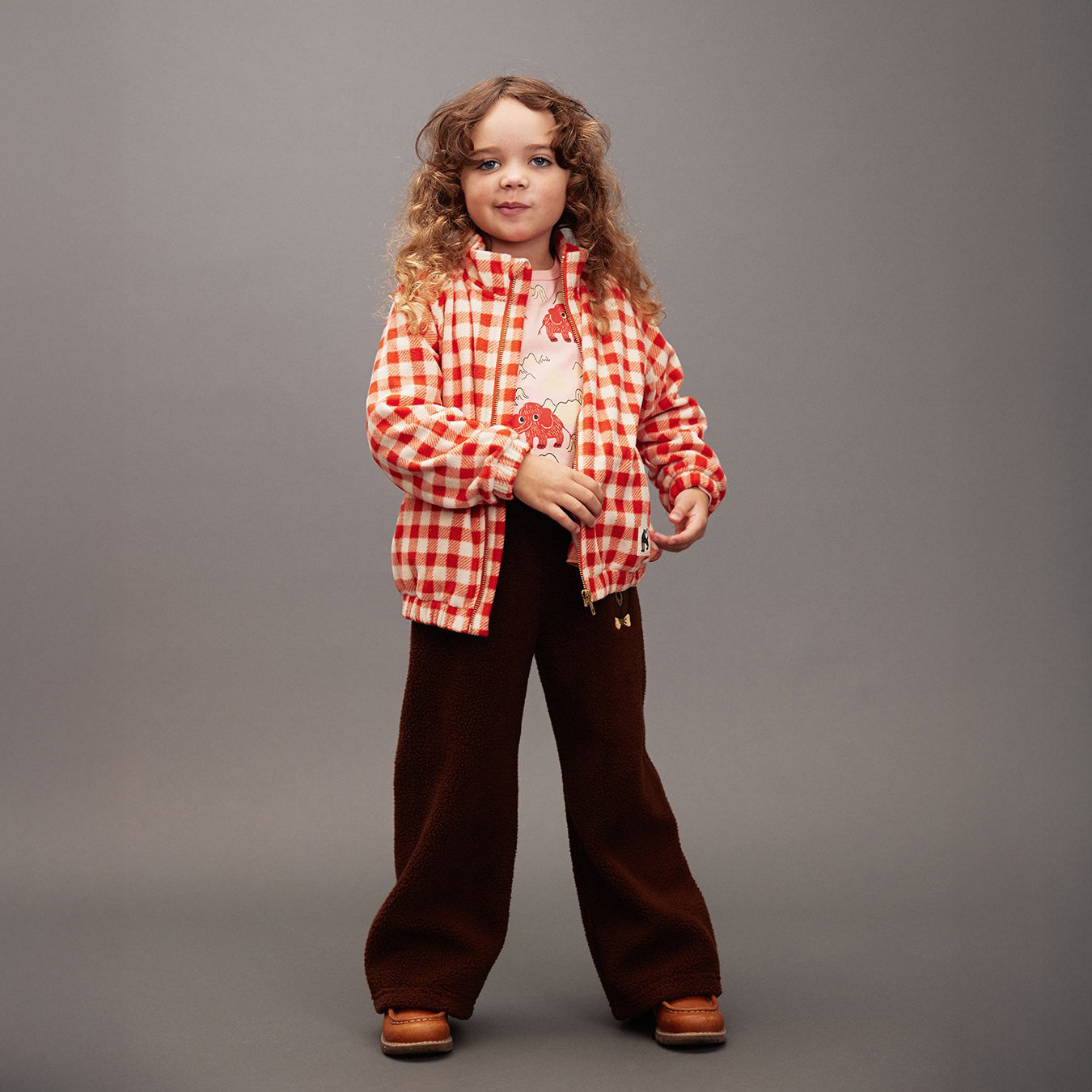 Child wearing a red checkered shirt and brown pants on a gray background