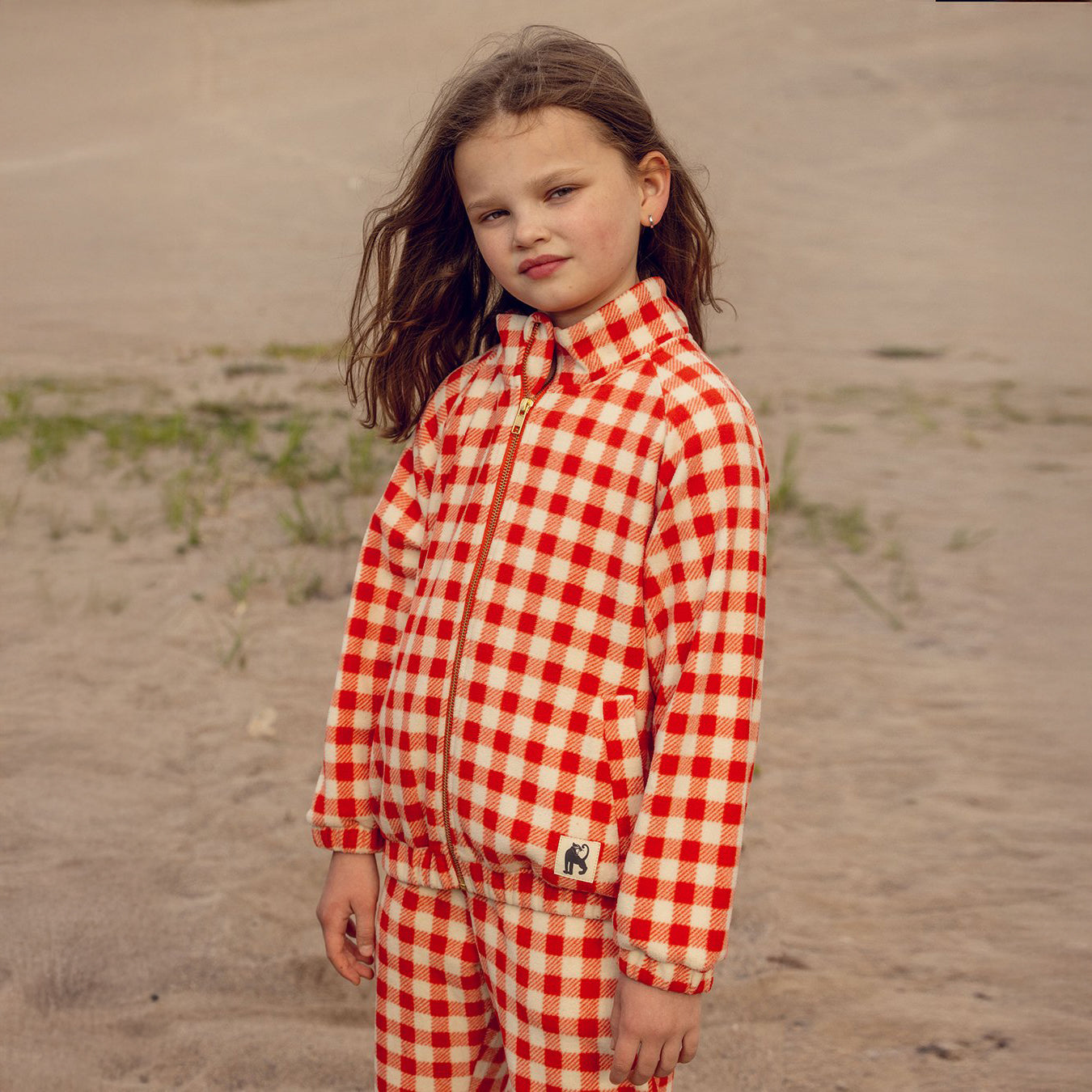 Young girl wearing a red and white checkered outfit standing on a sandy beach.