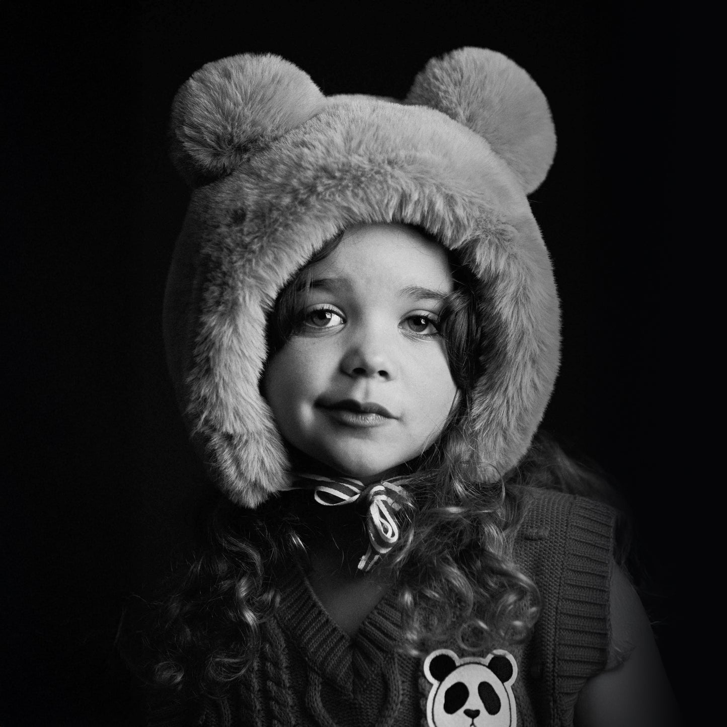 Black and white photo of a child wearing a fluffy bear hat with a dark background