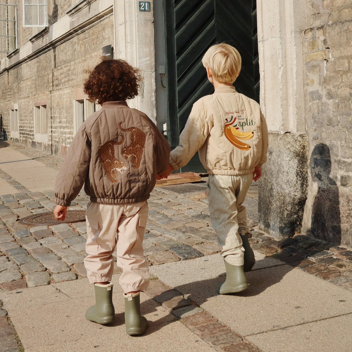 Two children walking hand in hand on a cobblestone street, wearing matching outfits with graphic designs.