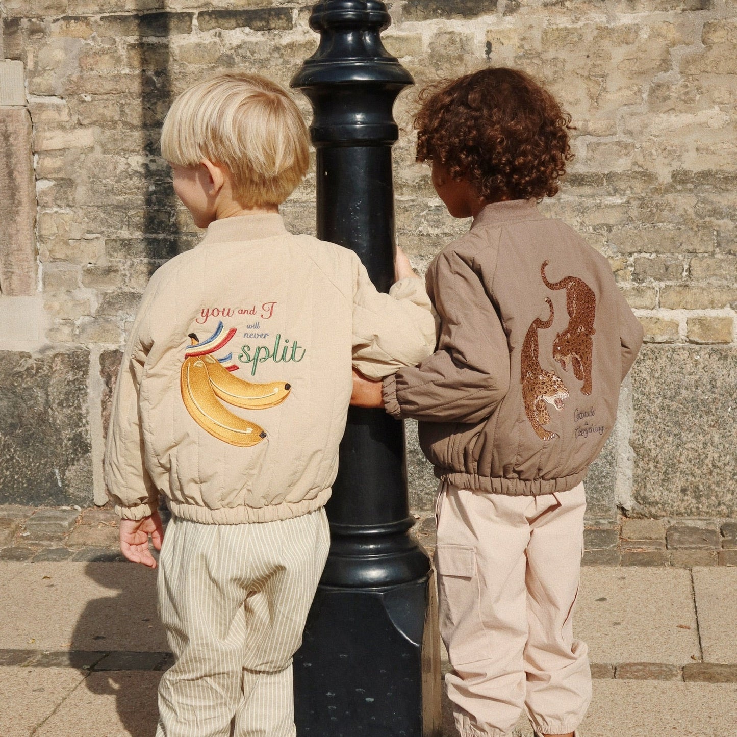 Two children wearing bomber jackets with animal prints standing next to a black lamp post.