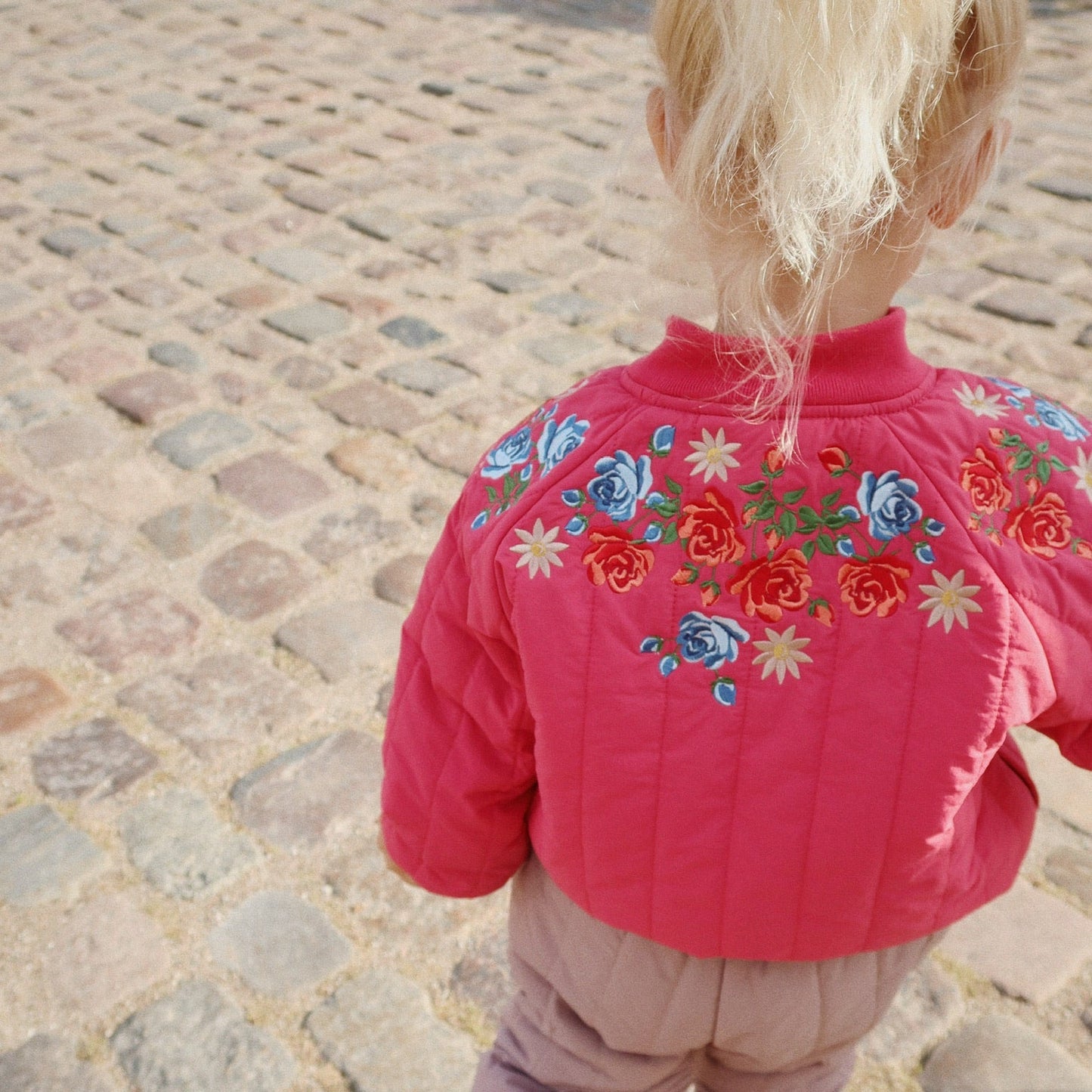 Child wearing a pink jacket with floral embroidery on a cobblestone street