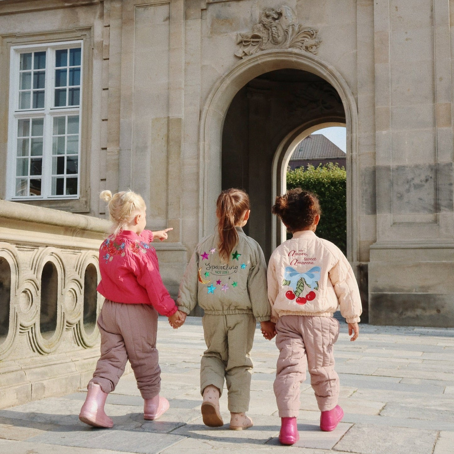 Three children in colorful outfits walking towards a stone archway.