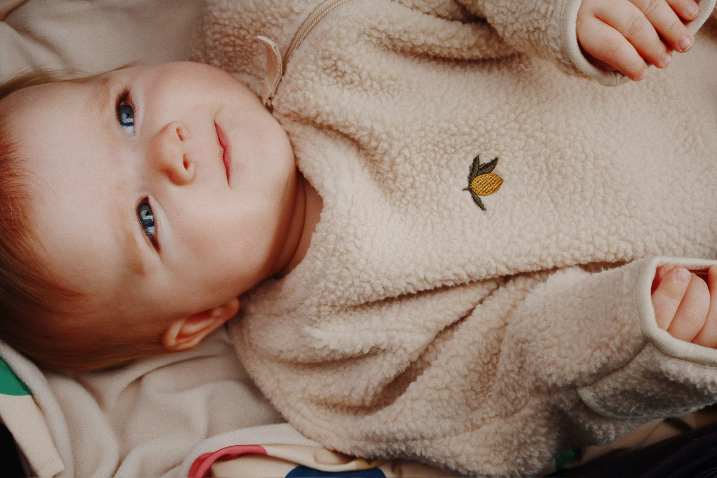 Baby wearing a beige fleece outfit with a small design, lying on a soft surface.
