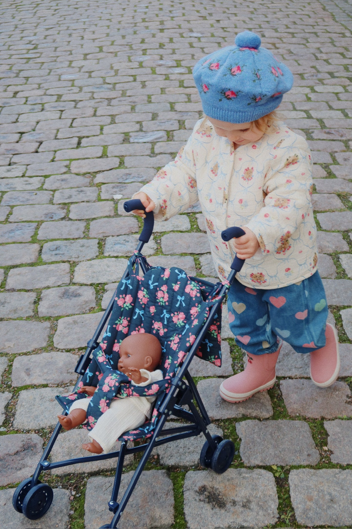 Child pushing a floral stroller with a doll on a cobblestone street
