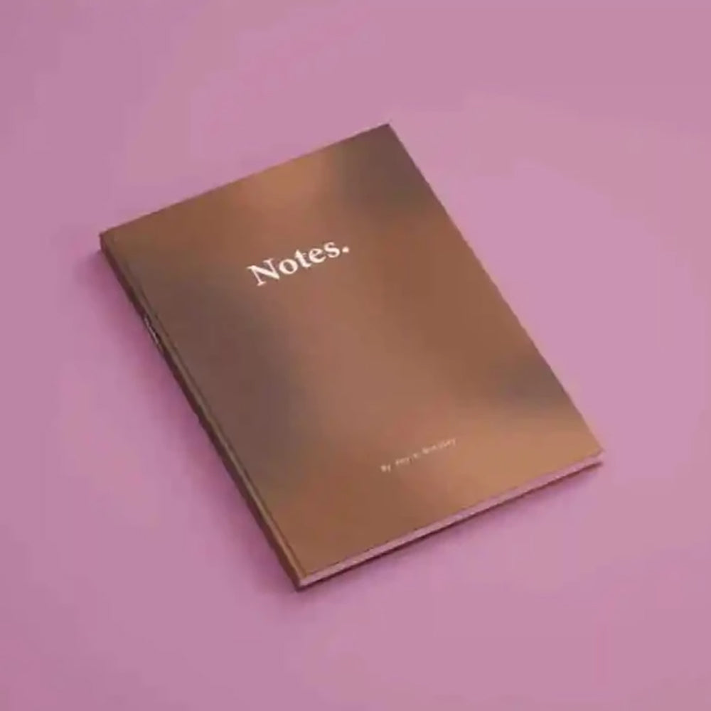 A bronze or copper colored book titled 'Notes...' placed on a pink surface.