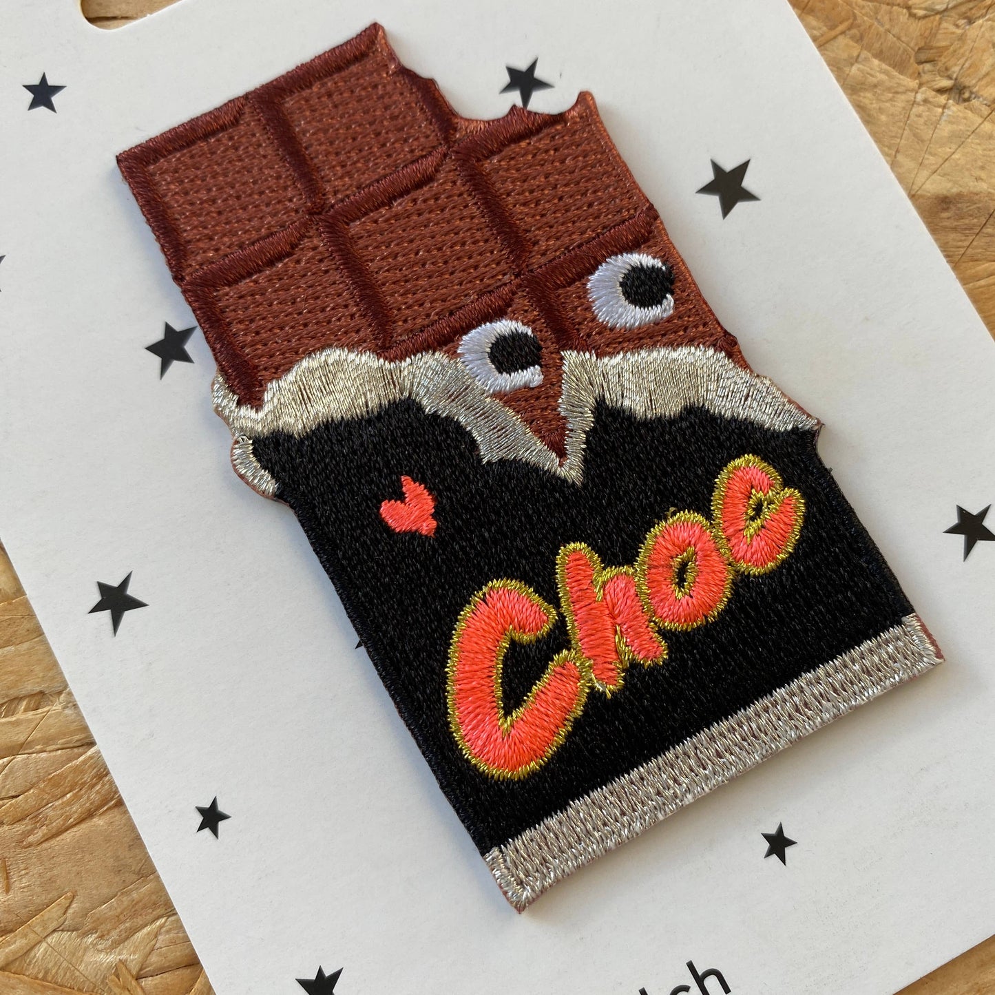A fun and quirky embroidered iron-on patch designed to look like a bitten chocolate bar. The patch features a rich brown chocolate section with raised details, googly-style embroidered eyes peeking out from the torn black-and-gold wrapper, and the word "Choc" stitched in bright orange and yellow with a tiny red heart.