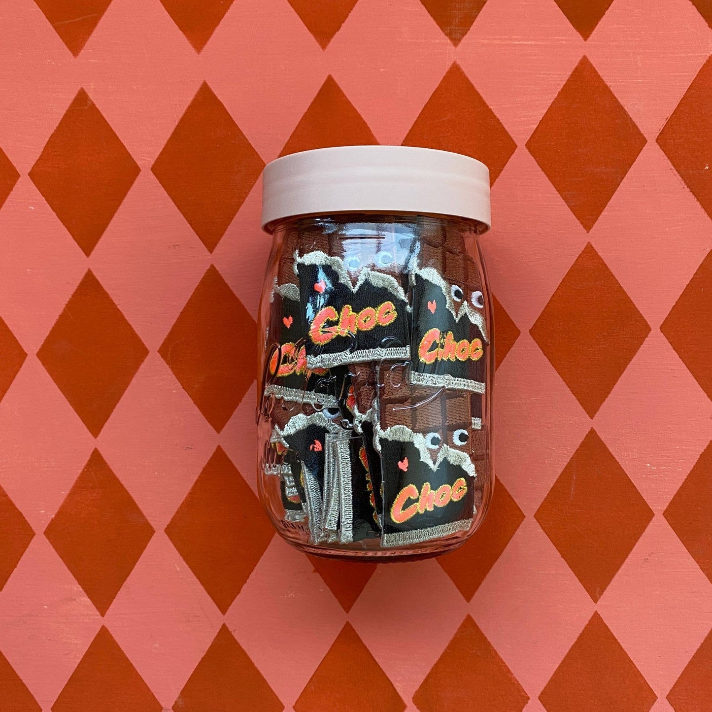 A clear glass jar filled with multiple Love Choc iron-on patches, placed against a bold retro-patterned background with red and pink diamond shapes. The patches inside the jar all feature the same playful chocolate bar design.