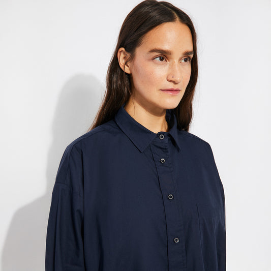 Close-up of the model in the oversized navy cotton shirt, focusing on the smooth, lightweight fabric and subtle sheen. The structured collar and neat black buttons contrast with the soft drape of the shirt.