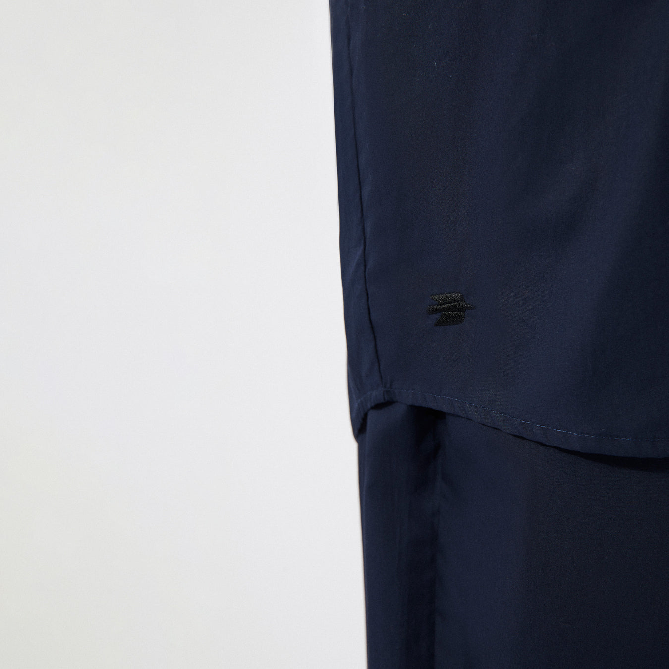 Close-up of the lower part of the navy oversized cotton shirt, focusing on a small, subtly embroidered black logo near the side seam. The fabric has a smooth, matte finish, and the hem is slightly curved.