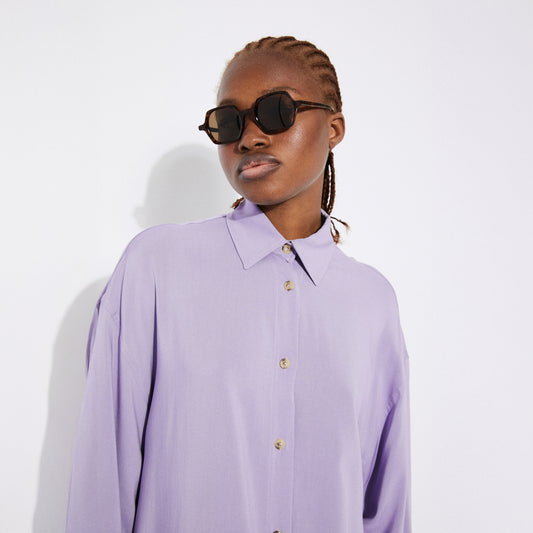Close-up of the model wearing the lilac button-up shirt, highlighting the pointed collar, button details, and soft bamboo fabric. The model wears black sunglasses and has neatly braided hair.