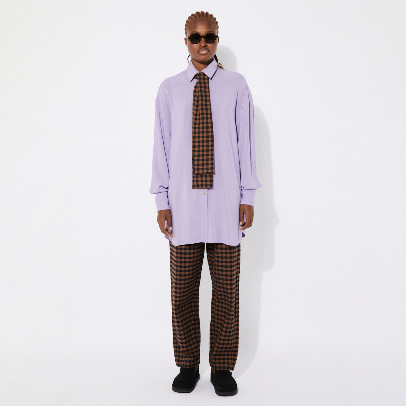 Model wearing the oversized lilac shirt styled with a matching brown gingham check tie. The tie drapes down the front, adding contrast to the soft lilac fabric. The model stands confidently, wearing black sunglasses.