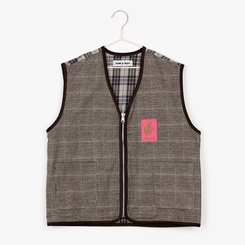 An image of a brown checkered/gingham zip up vest hanging up on a white hanger against the background of a white wall. The inside of the vest and the shoulders has a tartan pattern. To the right of the zip is a bright pink/coral patch that says Tom & Boy and had a cut-out of a donkey.