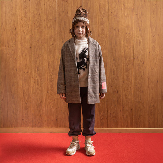 A white boy standing in front of a brown panelled wall in a room with red floors wearing am oversized gingham blazer, with a pink donkey on the arm of the blazer. Under the blazer is a cream knit jumper with a black donkey in the middle of it with pink outlining around it. This is paired with brown joggers with a pinstripe down the front, white socks and cream and light brown trainers. He also has on a brown and white knitted hat that has a sea pattern on it.