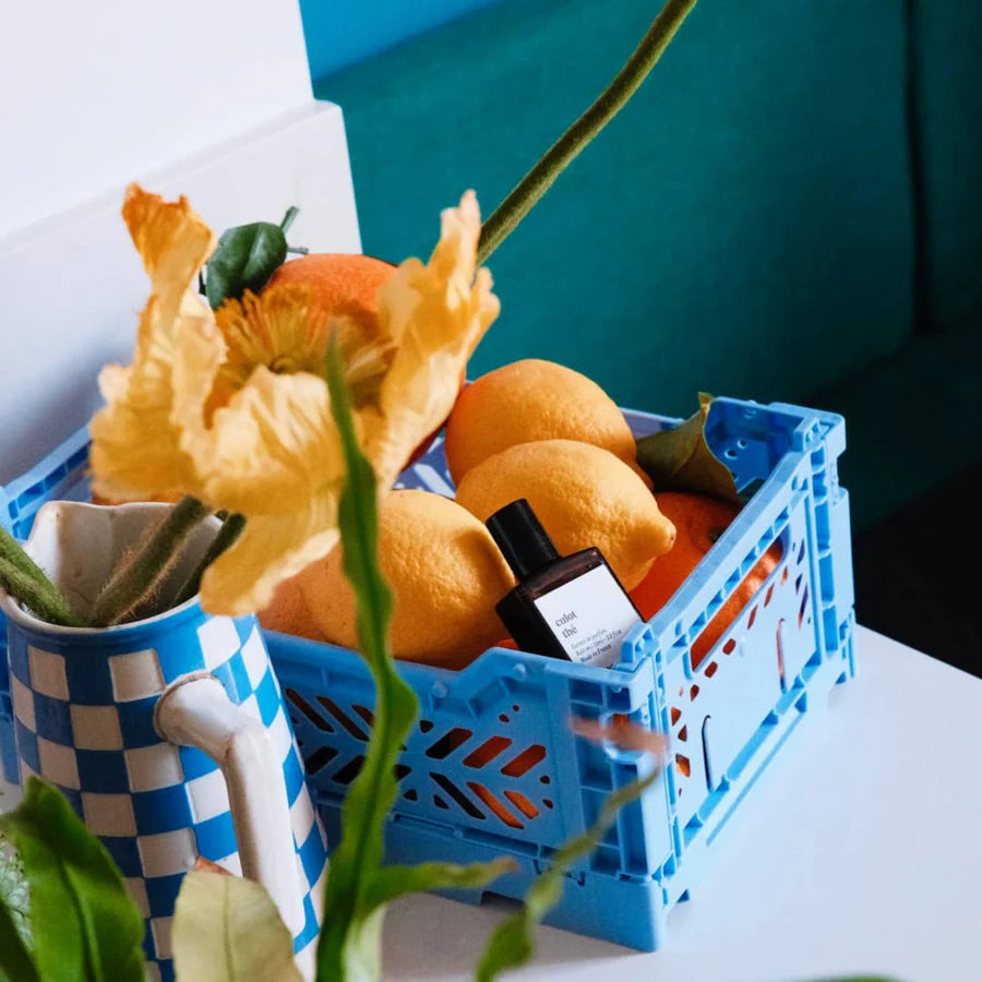 Blue crate with oranges, a bottle, and flowers on a white surface